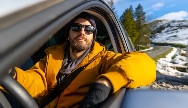 Un homme barbu portant des lunettes de soleil et un bonnet est assis au volant d'une voiture en hiver.