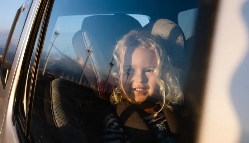 Une petite fille souriante aux cheveux blonds bouclés assise dans un siège auto, vue à travers la fenêtre.
