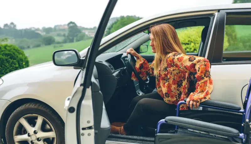 Une femme en robe à motifs floraux sort d'une voiture blanche, une fauteuil roulant à ses côtés.