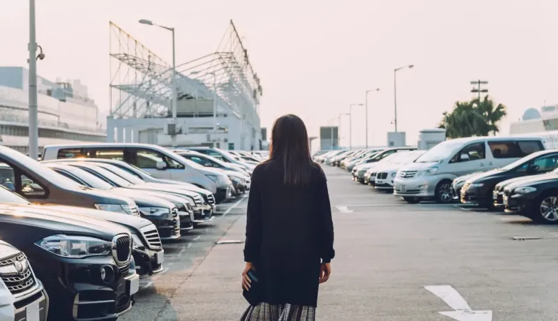 Vue de dos d'une femme seule marchant dans un grand parking rempli de voitures.