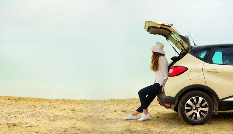 Une femme en chapeau assise sur le coffre ouvert d'une voiture, regardant un lac ou la mer.
