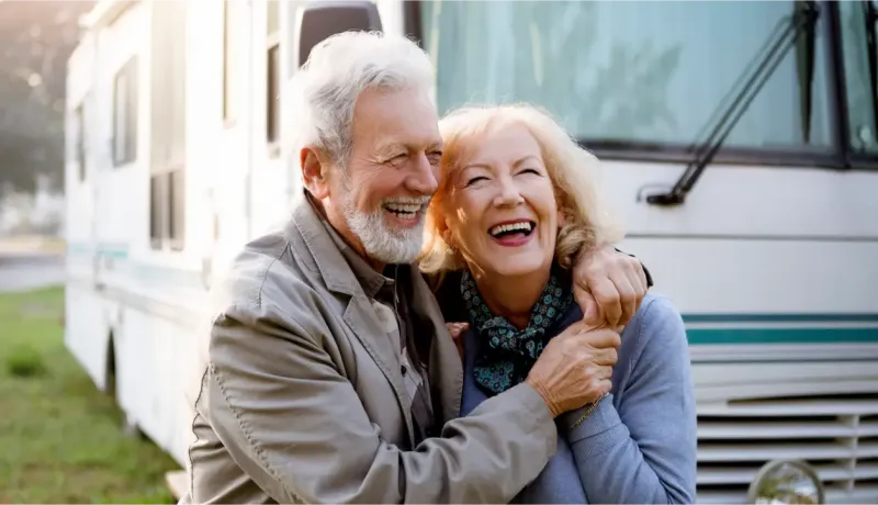 Un couple de personnes âgées souriantes et enlacées devant un camping-car, profitant de leur temps ensemble.