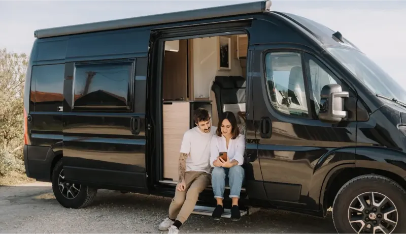 Un jeune couple assis sur le pas de la porte d'un van aménagé noir, utilisant un smartphone par une journée ensoleillée.