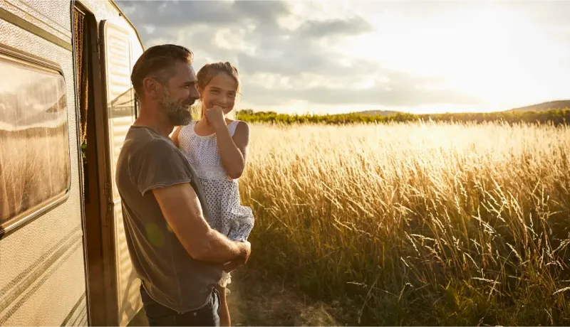 Un père souriant tient sa fille dans ses bras devant un camping-car, avec un champ de blé doré en arrière-plan.