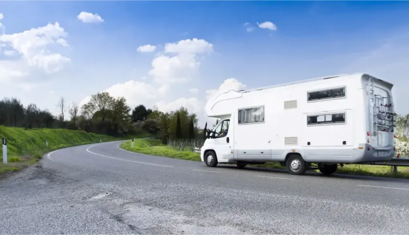Un grand camping-car blanc roule sur une route sinueuse à travers la campagne, sous un ciel bleu parsemé de nuages blancs.