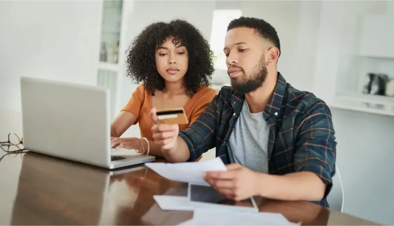 Un couple est assis à une table devant un ordinateur portable, la femme tenant une carte de crédit pendant qu'ils gèrent leurs finances.