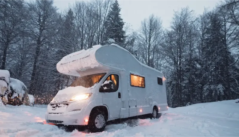 Un camping-car blanc enneigé est garé dans un paysage hivernal boisé, avec les lumières allumées créant une atmosphère chaleureuse.
