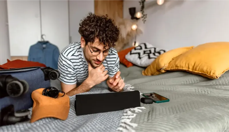 Un jeune homme aux cheveux bouclés et aux lunettes est allongé sur un lit, absorbé par le contenu d'une tablette ou d'un écran.