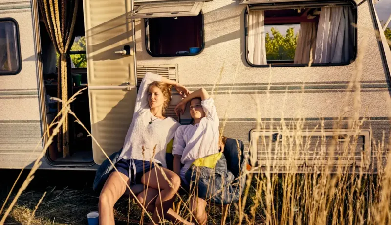 Deux personnes se détendent à l'extérieur d'une caravane par une journée ensoleillée, entourées d'herbes hautes et dorées.