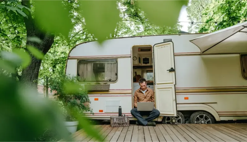 Un homme travaille sur son ordinateur portable, assis sur une terrasse en bois devant sa caravane entourée d'arbres verdoyants.