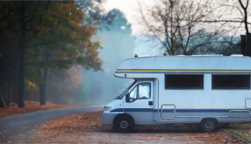 Un camping-car blanc avec des rayures bleues est garé sur le côté d'une route de campagne, au milieu de feuilles d'automne et d'arbres dépouillés.