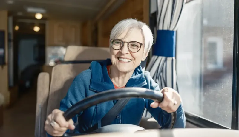 Une femme souriante, les cheveux gris, vêtue d'un pull à capuche bleu, est assise au volant de son camping-car, prête à prendre la route.