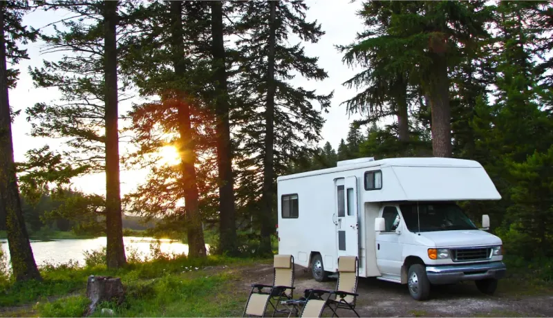 Un camping-car blanc est garé au bord d'un lac, entouré de grands pins, avec le soleil couchant en arrière-plan.