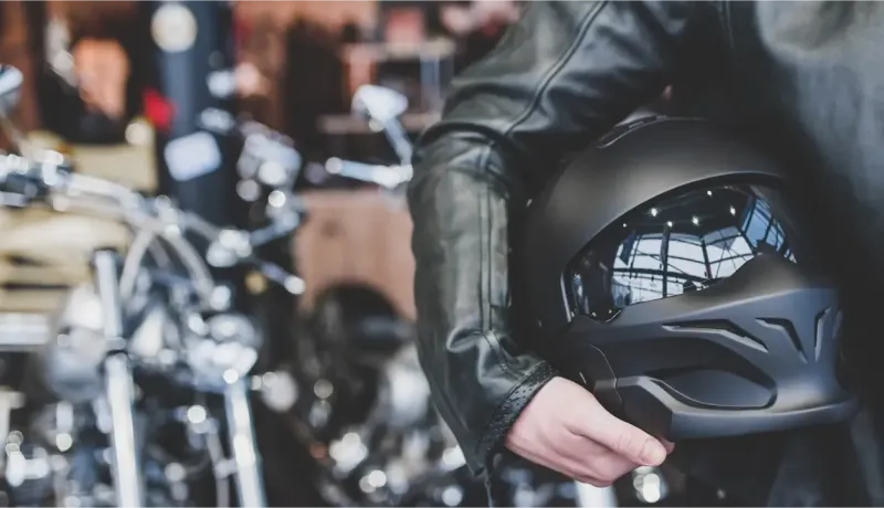 Un motard tenant un casque noir mat avec une visière réfléchissante, prêt à l'aventure devant une moto.