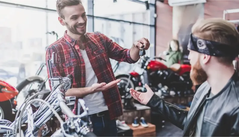 Un vendeur souriant remet les clés d'une moto à un client barbu et tatoué dans un magasin de motos.