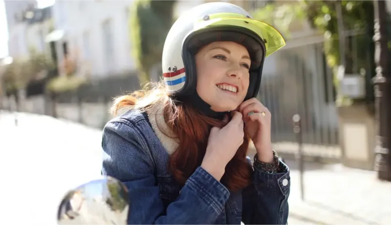 Une jeune femme rousse souriante ajuste son casque de moto vintage blanc et coloré, prête à partir.