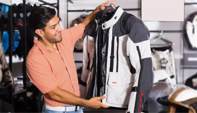 Un homme souriant tient une veste de moto blanche et grise sur un cintre, l'inspectant dans un magasin d'équipement.