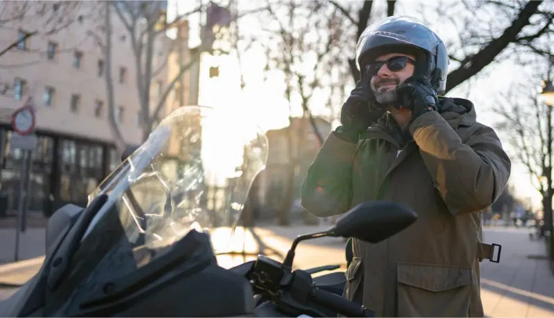 Un motard ajuste son casque et ses lunettes de soleil sur une moto, avec un soleil éclatant en arrière-plan urbain.