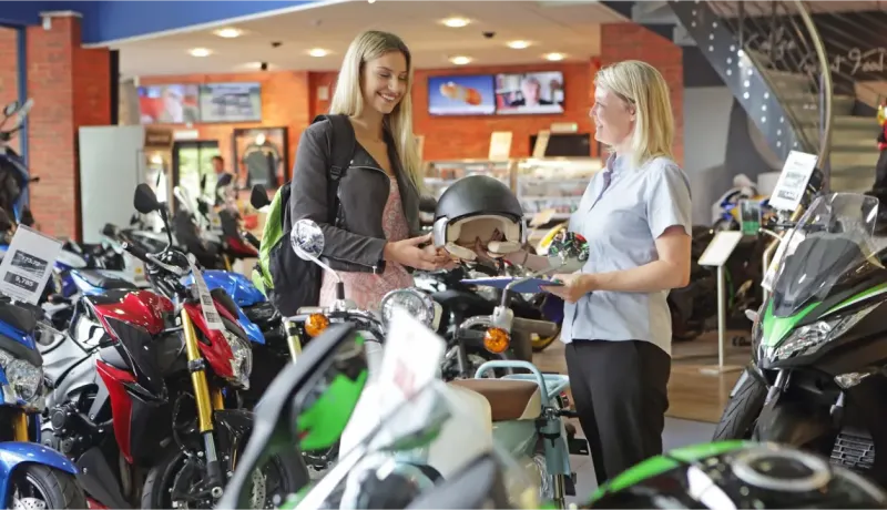 Une jeune femme reçoit un casque de moto d'une vendeuse souriante dans un magasin de motos.