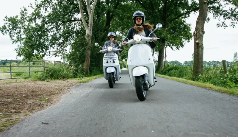 Deux personnes en casque conduisant des scooters de type Vespa sur une route de campagne bordée d'arbres.