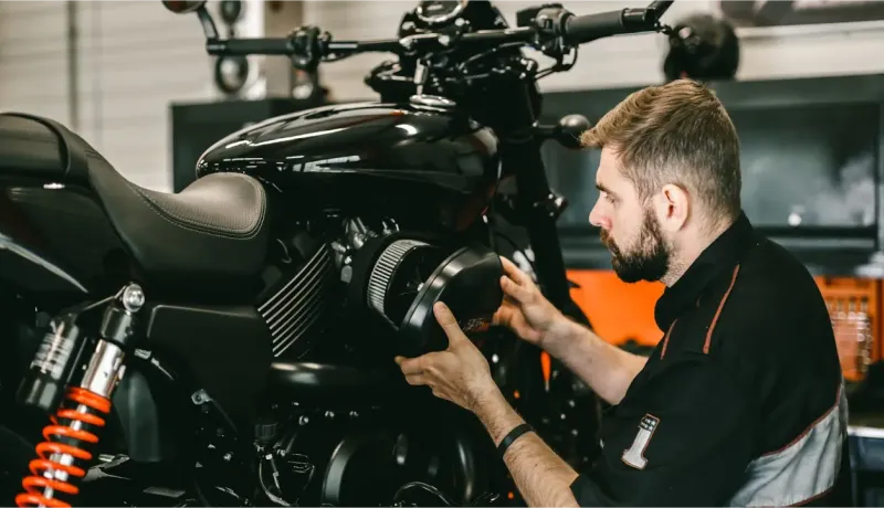 Un homme barbu, mécanicien, travaille sur le moteur d'une moto noire en atelier.