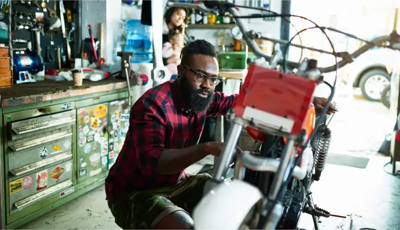 Un homme barbu avec des lunettes travaille sur une moto dans un garage encombré d'outils et de pièces.