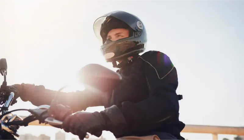 Un motard en tenue de protection et casque intégral regarde vers le ciel sous un soleil éclatant.