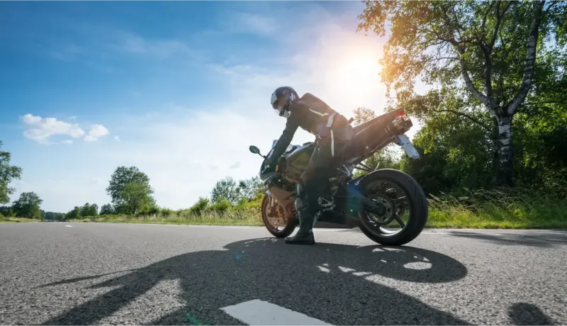 Un motard se penche sur sa moto sportive sur une route de campagne ensoleillée, sous un ciel bleu.