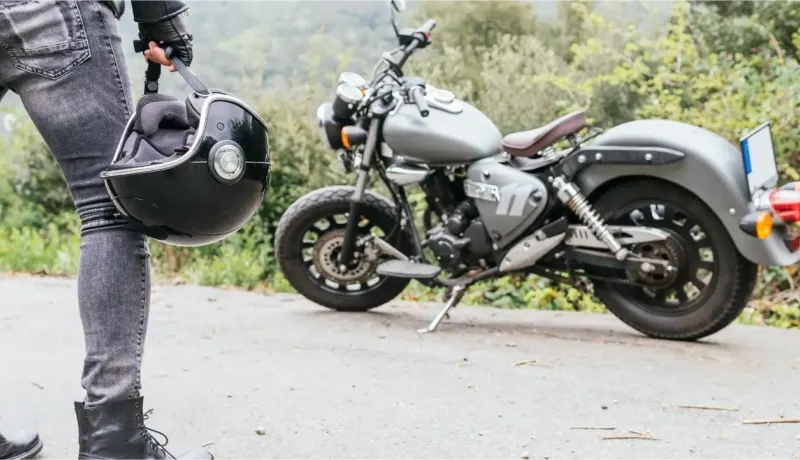 Un motard tenant son casque noir à côté d'une moto custom grise garée sur le côté d'une route.