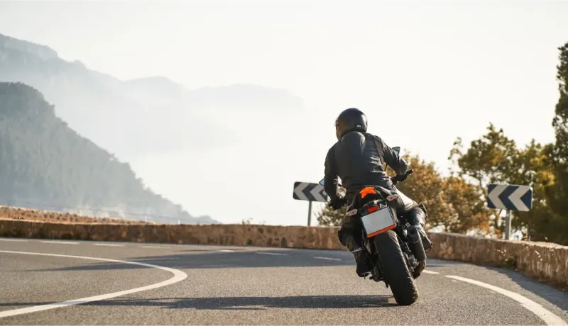 Vue arrière d'un motard prenant un virage sur une route de montagne sinueuse, avec des paysages brumeux.