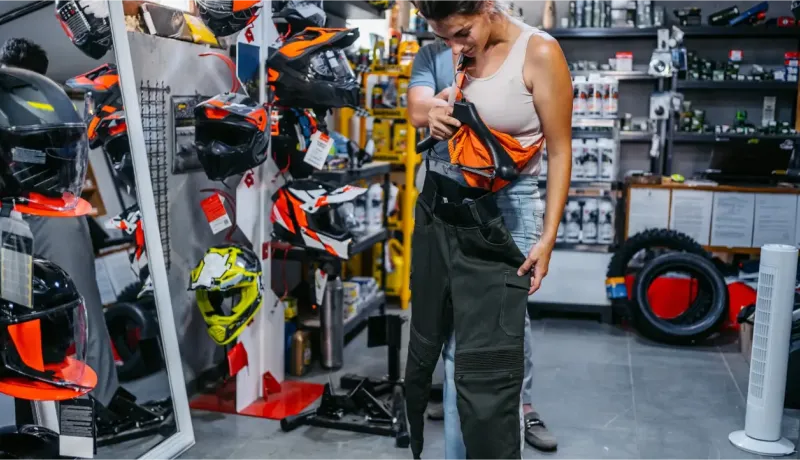 Une femme essaie un pantalon de moto dans un magasin rempli de casques et d'équipements.