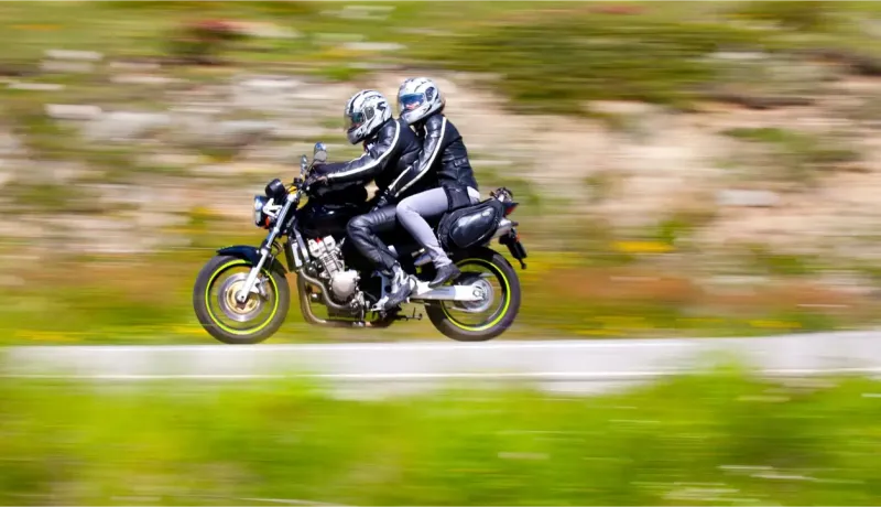 Deux motards en casque et blousons de cuir roulent à vive allure sur une route de campagne verte.