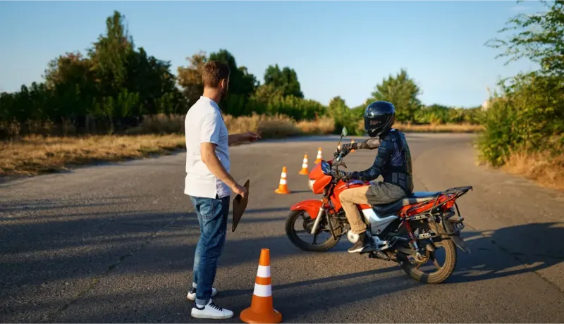 Un instructeur guide un élève motard en casque et équipement sur une moto rouge lors d'un cours.