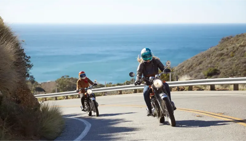 Deux femmes motardes sur leurs motos roulant sur une route côtière ensoleillée avec l'océan en fond.