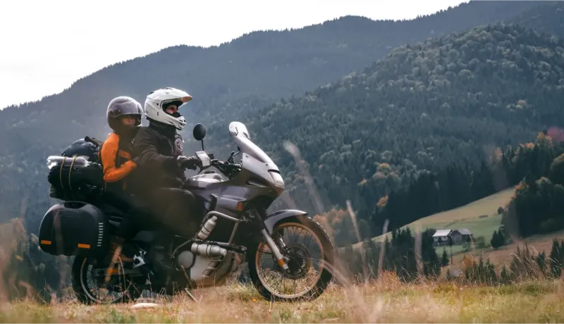 Deux motards avec bagages sur une moto de tourisme dans un paysage montagneux verdoyant et vallonné.