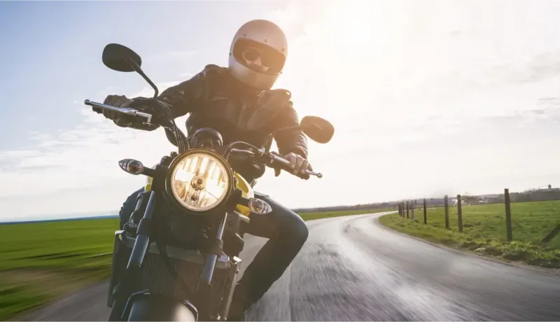 Un motard en blouson de cuir et casque roule sur une route sinueuse en campagne, au soleil couchant.