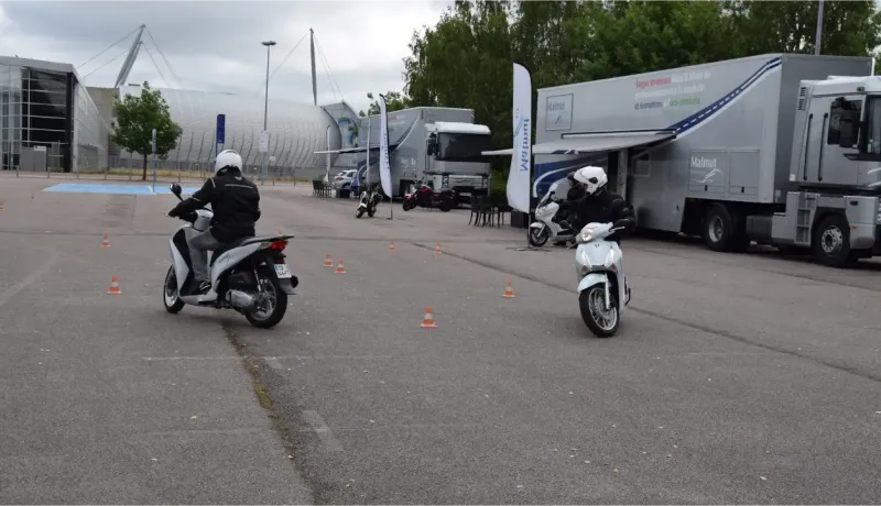 Des élèves motards en scooter pratiquent la conduite sur un parking avec des cônes, près de camions.