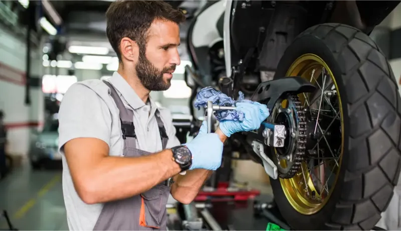 Un mécanicien barbu en combinaison grise et gants bleus travaille sur la roue arrière d'une moto.