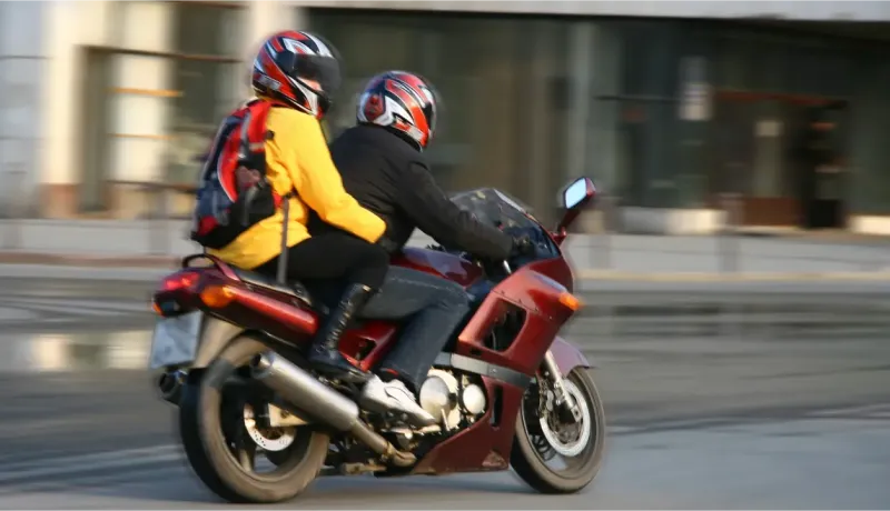 Un couple en casque de moto sur une moto rouge en ville. L'homme conduit, et la femme, avec un sac à dos, est assise derrière lui.