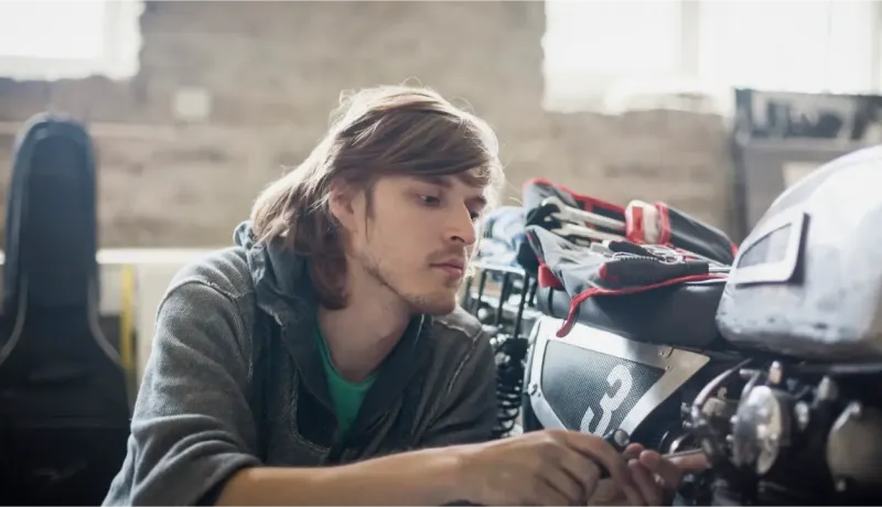 Un jeune homme aux cheveux longs travaille méticuleusement sur les détails d'une moto dans un garage.