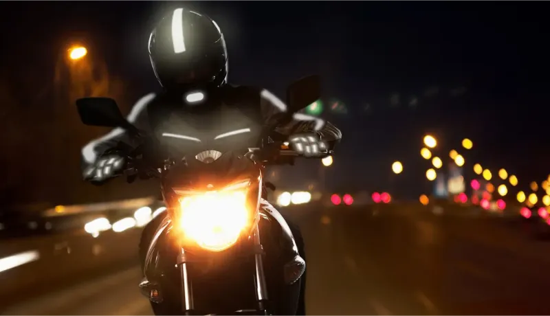 Un motard sur sa moto la nuit, vêtu d'un équipement réfléchissant, avec le phare allumé.