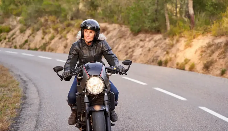Une femme mature en blouson de cuir et casque noir conduit une moto sur une route de campagne.