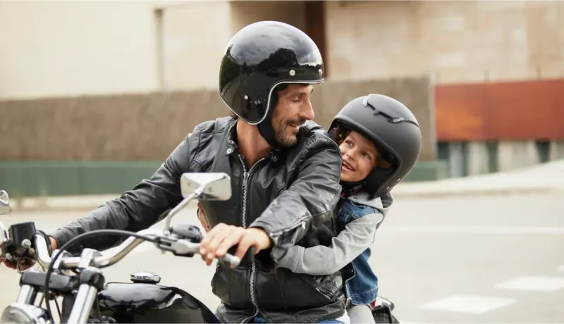 Un père souriant et son enfant en casque noir sur une moto, partageant un moment de complicité.