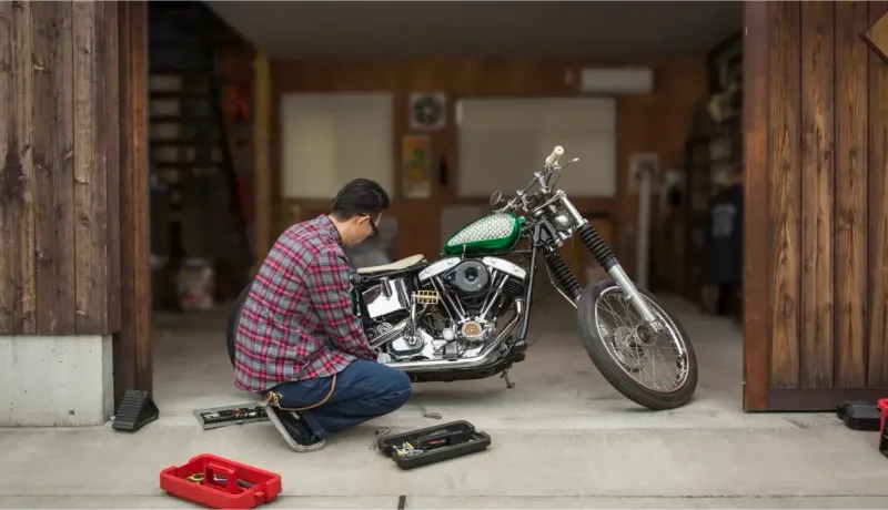 Un homme accroupi répare une moto custom dans un garage en bois ouvert avec des outils.