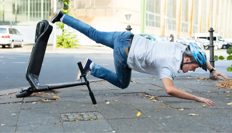 Un homme en jean et t-shirt avec un casque bleu tombe à côté de sa trottinette électrique sur un trottoir.