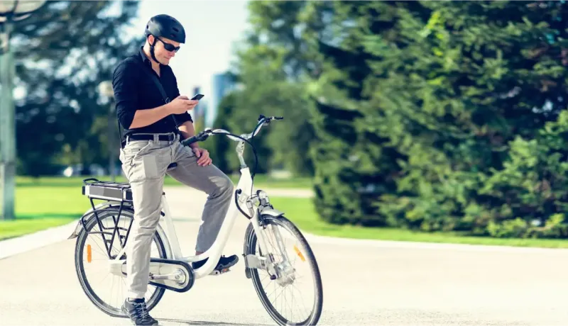 Un homme casqué et lunettes de soleil, assis sur un vélo électrique blanc, regarde son téléphone.