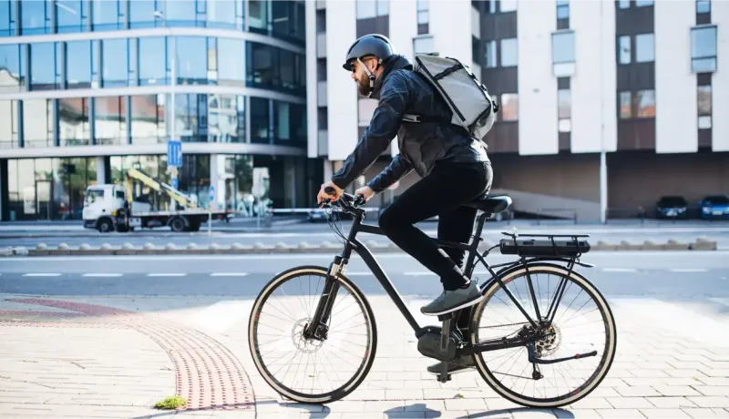 Un homme en casque et sac à dos roule sur un vélo électrique noir en ville, avec des bâtiments modernes en arrière-plan.