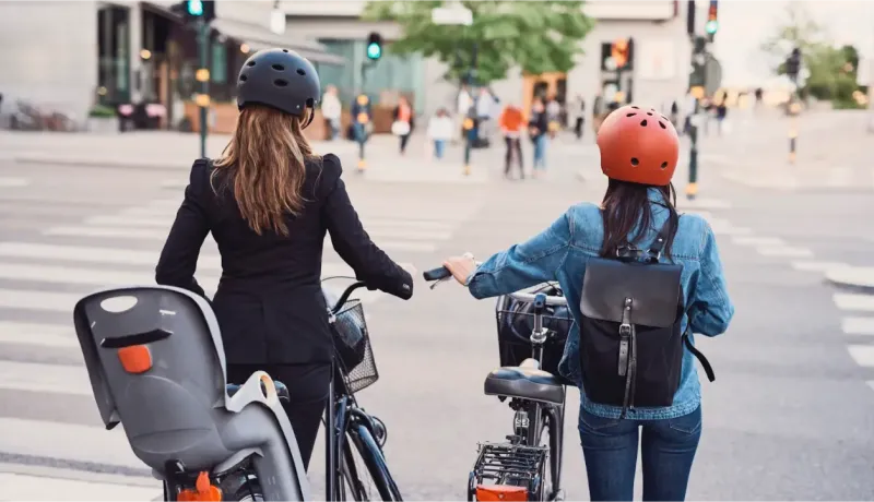 Deux femmes avec des casques sur la tête, l'une avec un siège enfant, l'autre avec un sac à dos, traversent la rue à vélo.