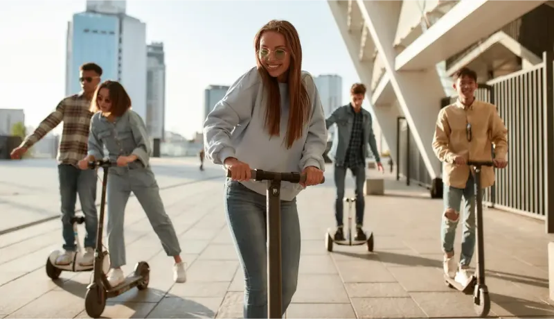 Un groupe de jeunes souriants s'amuse en trottinette et en hoverboard sur un large chemin extérieur.
