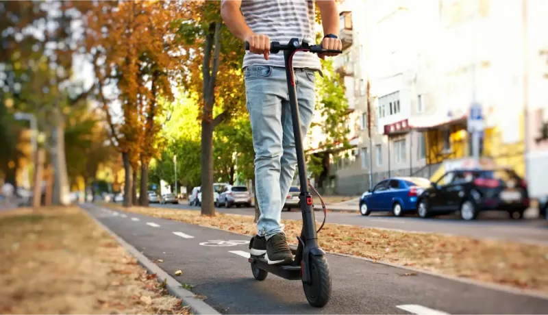 Un homme roule sur une trottinette électrique sur une piste cyclable bordée d'arbres en automne.